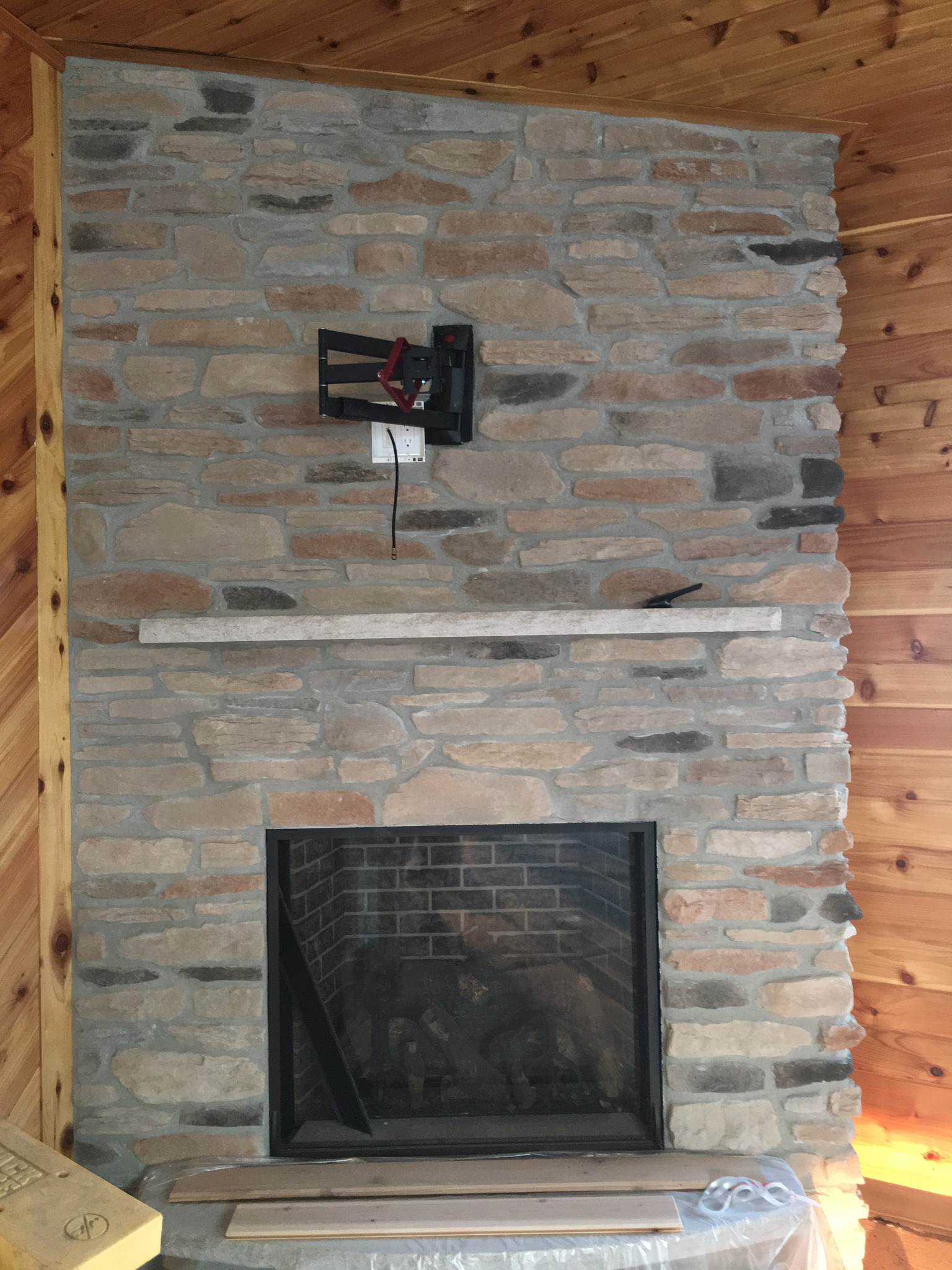 A stone fireplace with a black metal fireguard in a room with wooden walls and ceiling. There are some wooden pieces and a plastic bag on the mantel and floor.