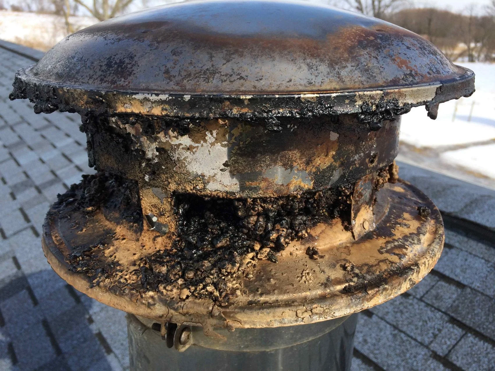Close-up of a rusty metal object with charred and burnt residue, possibly part of a chimney cap or vent, against a backdrop of snow and trees.
