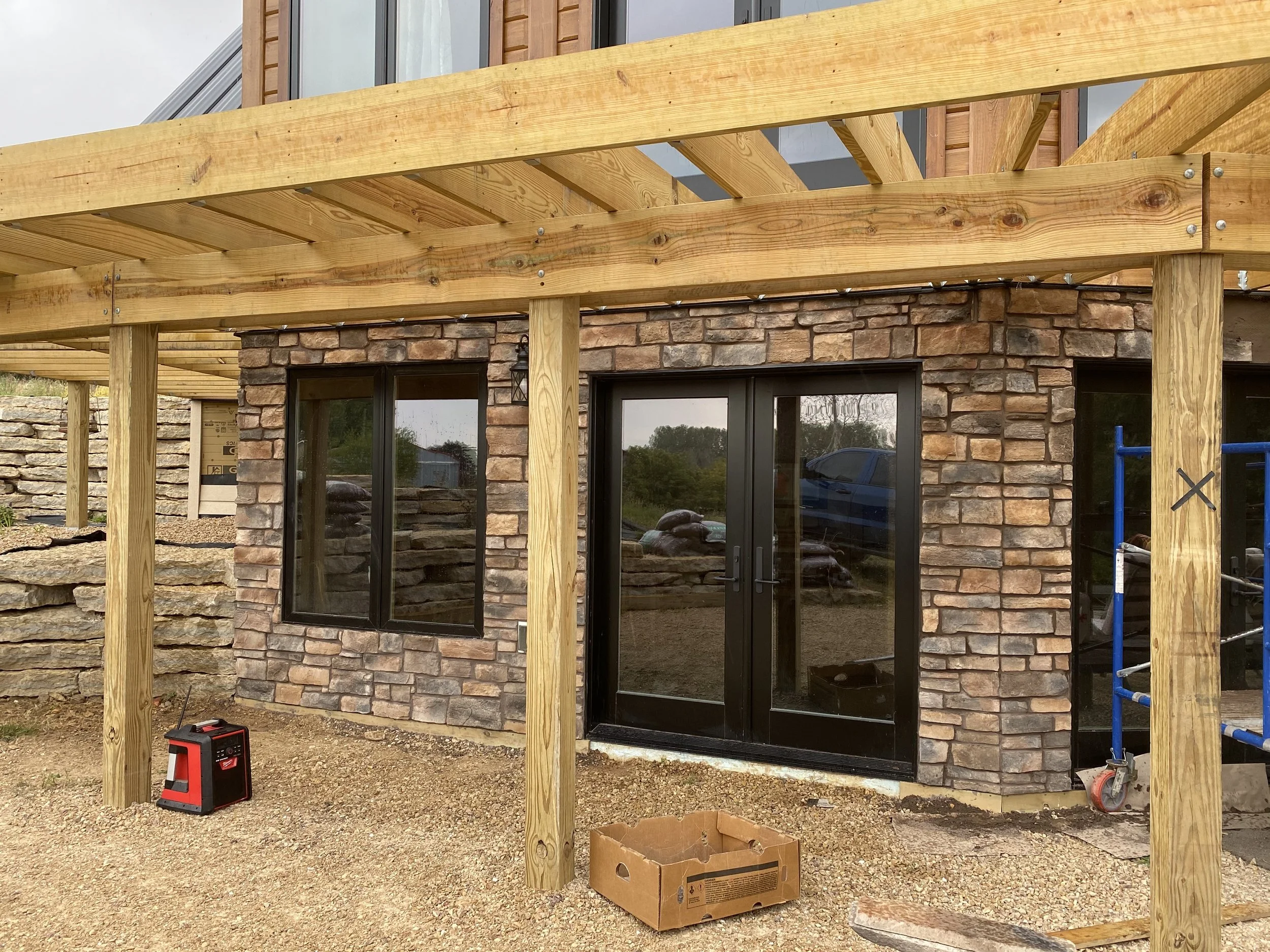 Construction site showing a partially built wooden deck attached to a stone building with large glass doors. The deck's wooden beams and planks are visible, with some tools and construction materials on the ground.
