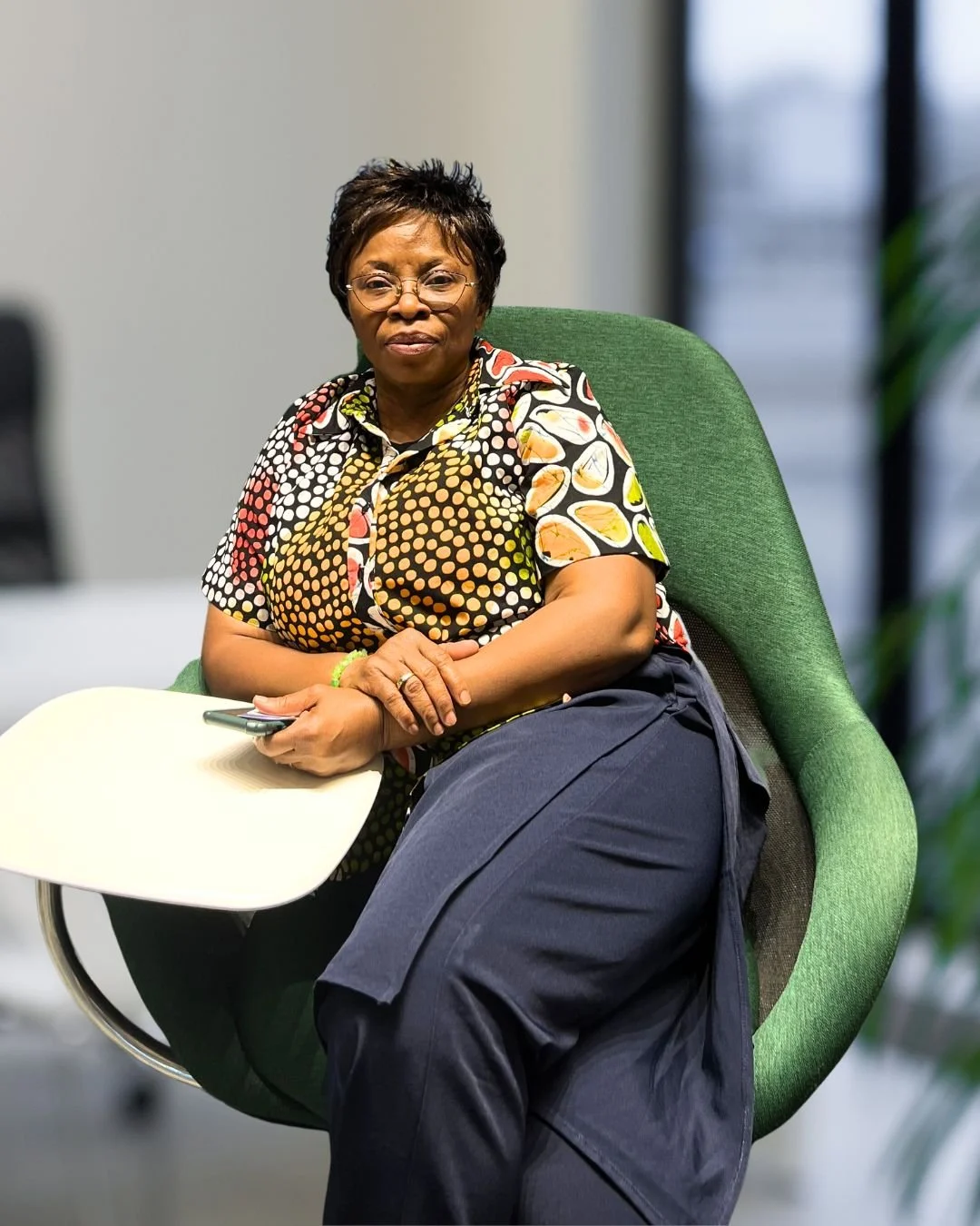 A middle-aged woman with short hair and glasses sitting in a green chair, wearing a colorful patterned shirt and dark pants, holding a smartphone in her left hand with a white tray on her lap.