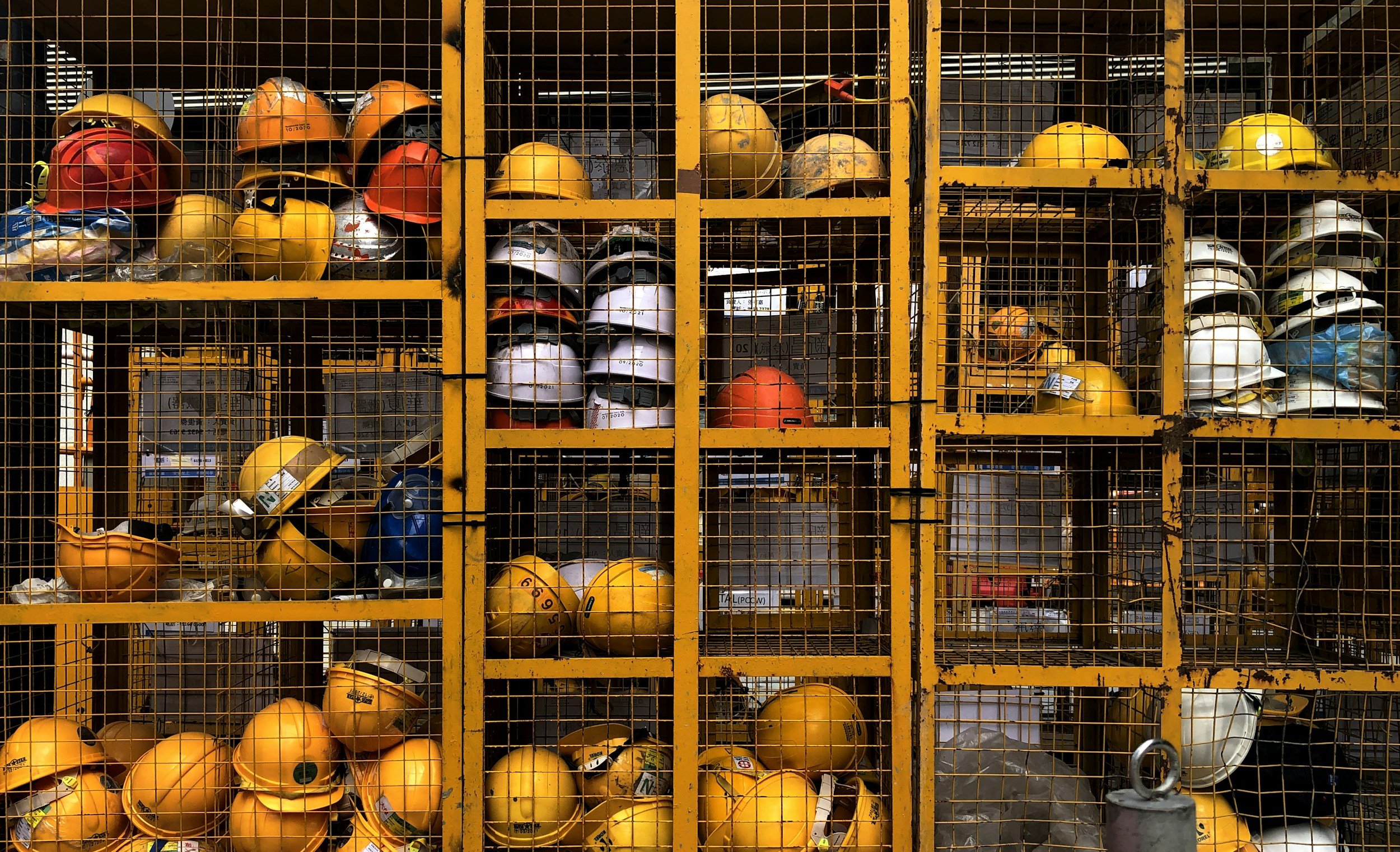 A large industrial shelving unit filled with various safety helmets in yellow, white, red, orange, and blue, behind a metal grid.
