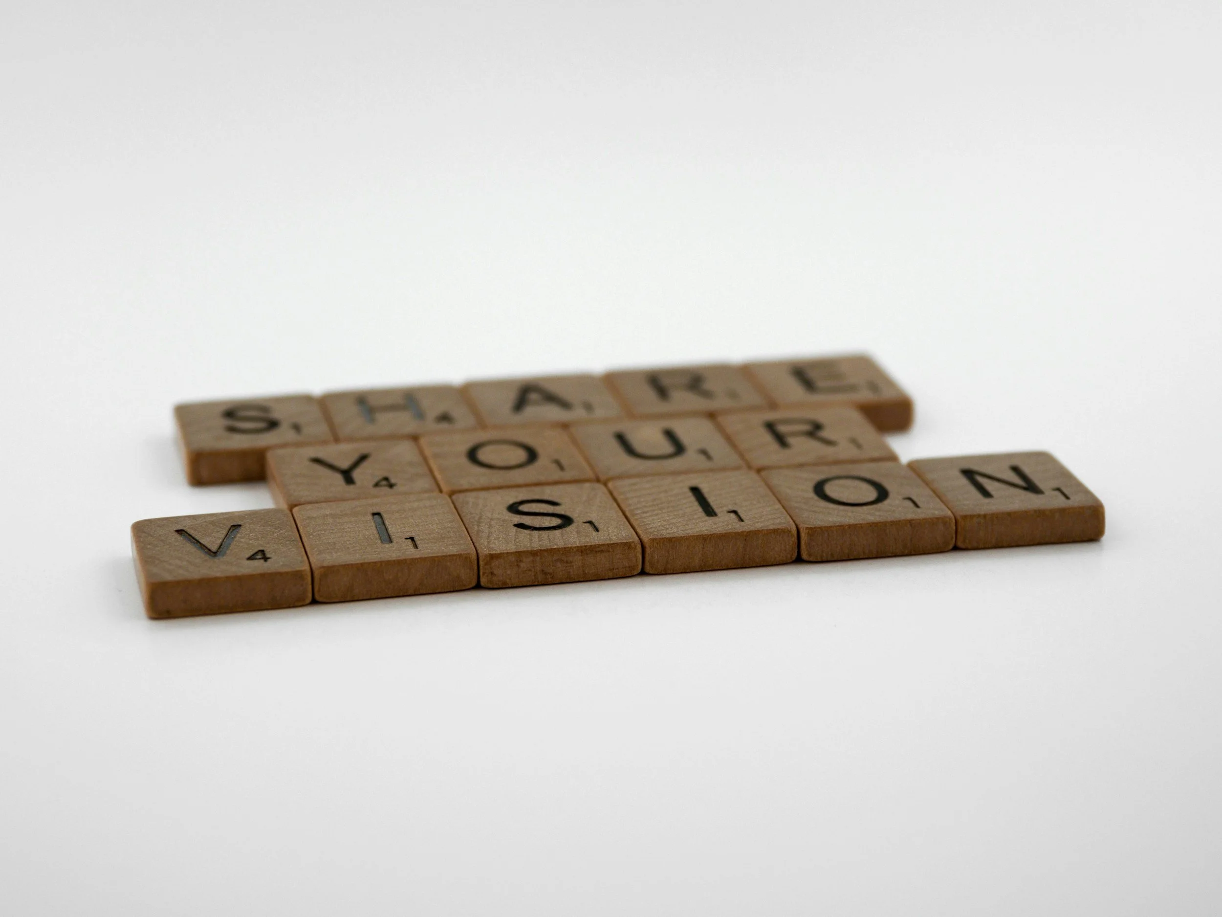 Wooden Scrabble tiles spell out the phrase "Share Your Vision" on a white background.