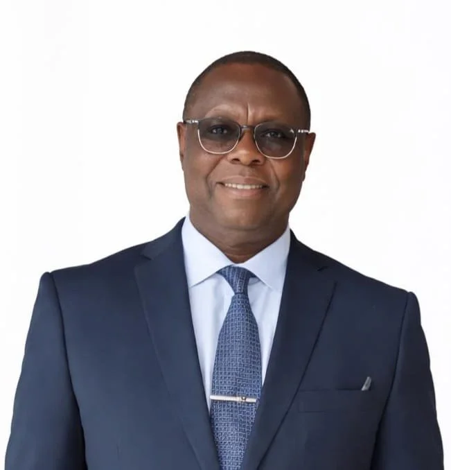 A smiling man with dark skin wearing glasses, a navy blue suit, a light blue shirt, and a patterned blue tie, standing against a plain white background.