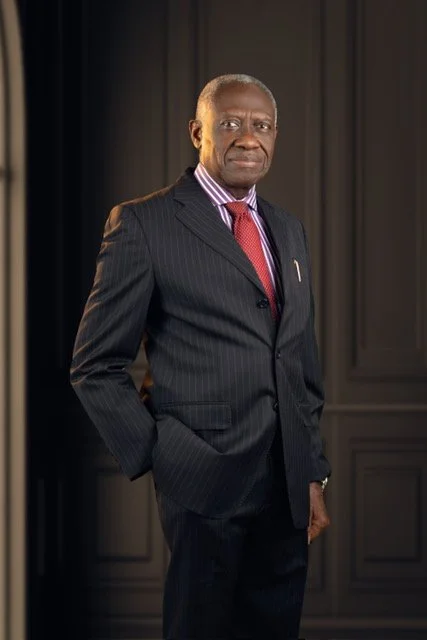 An older man wearing a dark pinstripe suit, pink and white striped shirt, and red tie, standing in front of a dark background with paneled walls.
