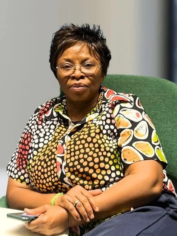 An older woman with short black hair, wearing glasses, a colorful patterned shirt, and a green wristband, sitting on a green chair, holding a smartphone in her right hand, in a well-lit indoor setting.