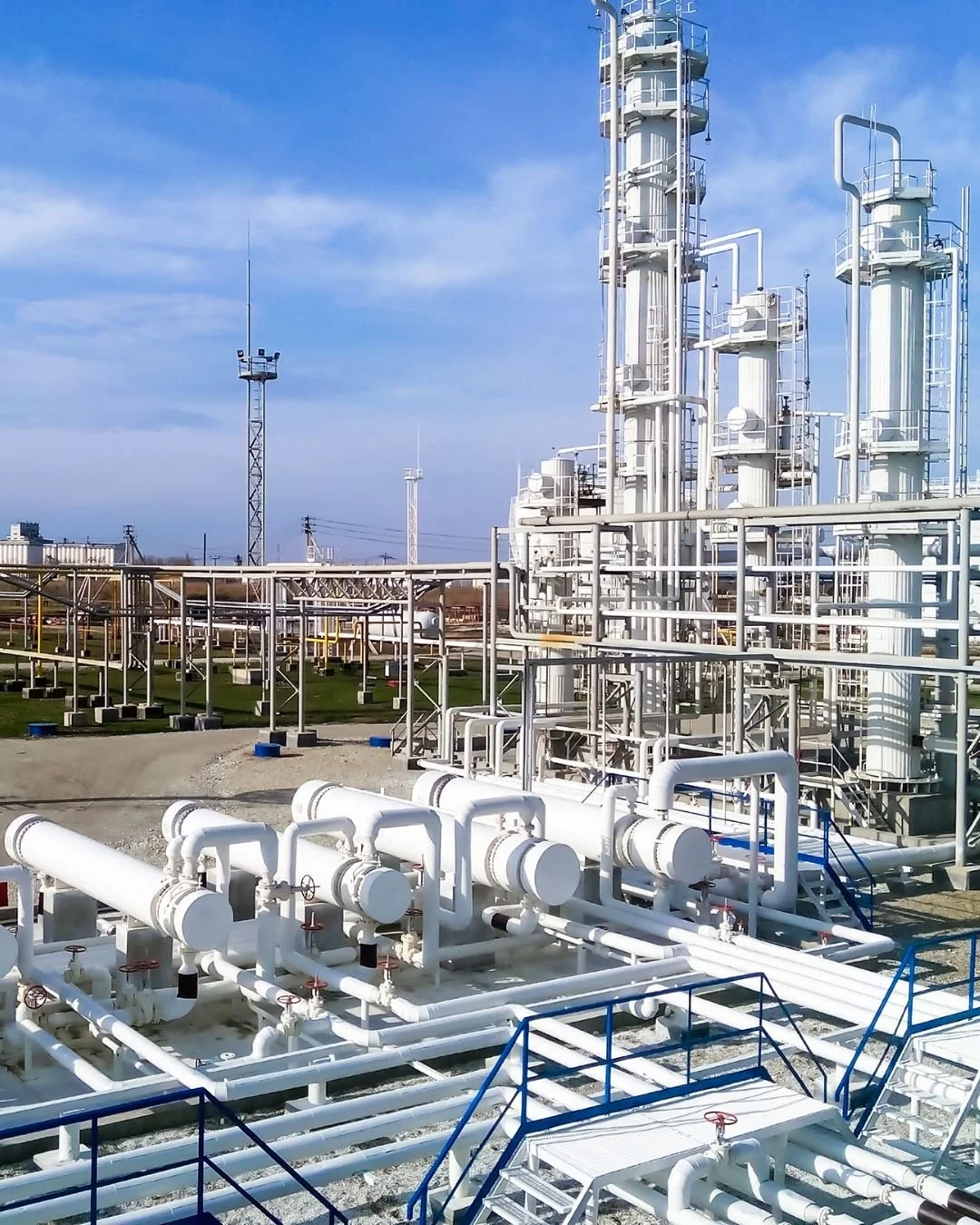 Industrial facility with large white pipes, tanks, and metal structures against a blue sky.
