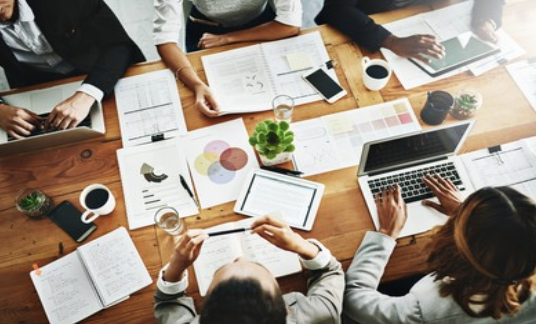 A top-down view of a collaborative work meeting with five people at a wooden table. The table is filled with laptops, tablets, papers, notebooks, and color swatches. There are plants, coffee cups, water glasses, and smartphones. The individuals are engaged with their devices and documents.