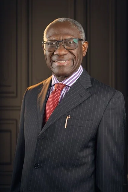 An older African American man wearing a dark pinstripe suit, a light purple striped shirt, a red tie, and glasses, smiling at the camera against a dark background.