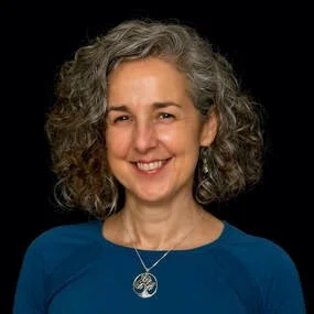 MYA Secretary, Mary Beth Thomas, smiling, wearing a blue top and a pendant necklace, against a black background