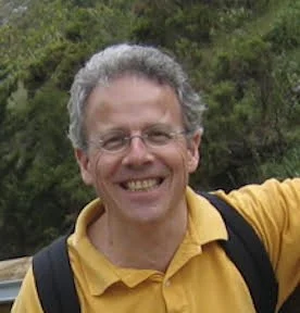 MYA member, John Brady, smiling outdoors, wearing glasses, a yellow shirt and a backpack