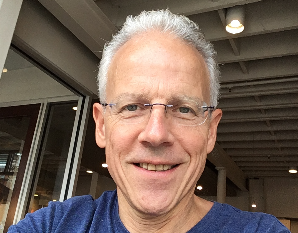 MYA member, John Brady, smiling outdoors, wearing glasses, a yellow shirt and a backpack