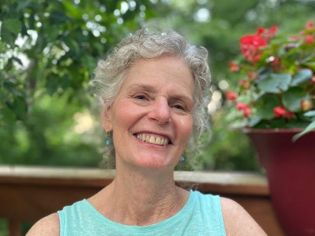 MYA board member at large, Ann Ross, in an outdoor setting, wearing a light blue sleeveless top and earrings, with green trees and pink flowers in the background