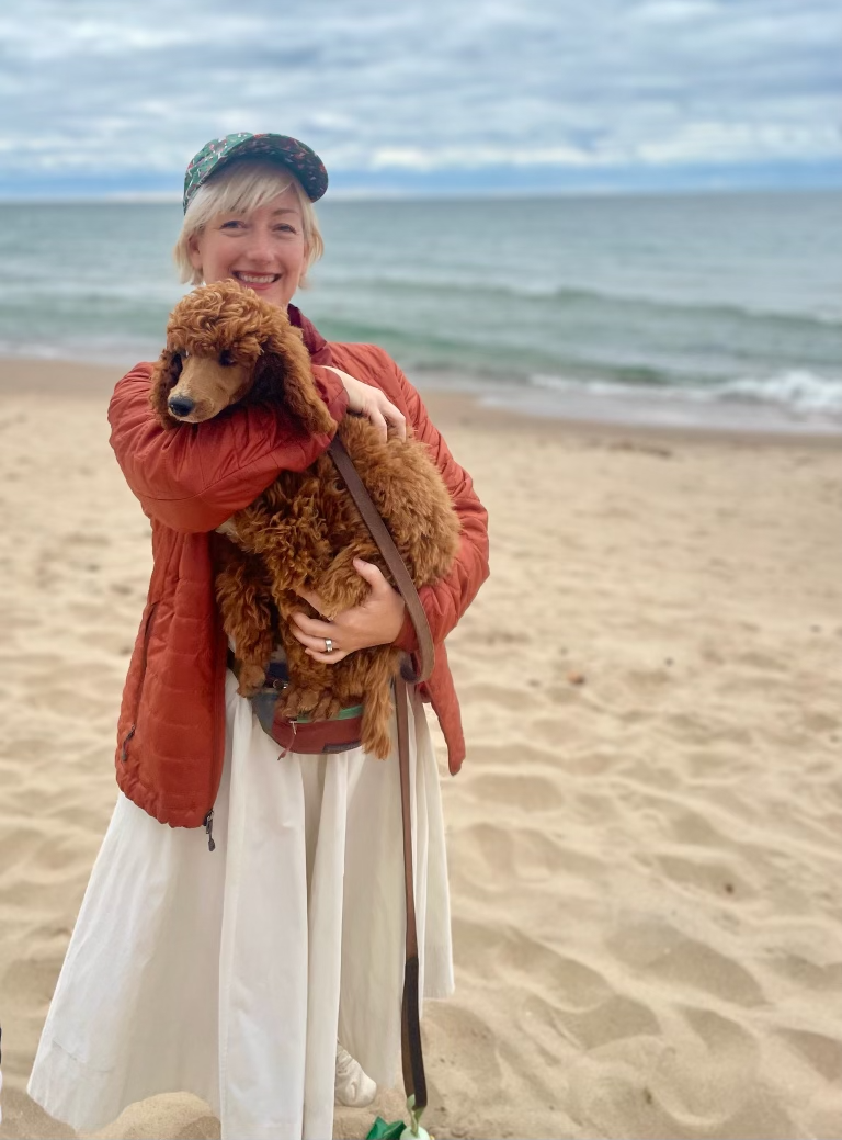MYA Vice President, Julie Klick, smiling at the camera while holding her curly-haired puppy on a sandy beach with the ocean and cloudy sky in the background