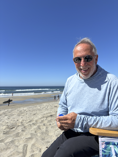 MYA President, Howard Dembs, with sunglasses sitting on the beach near the ocean on a sunny day
