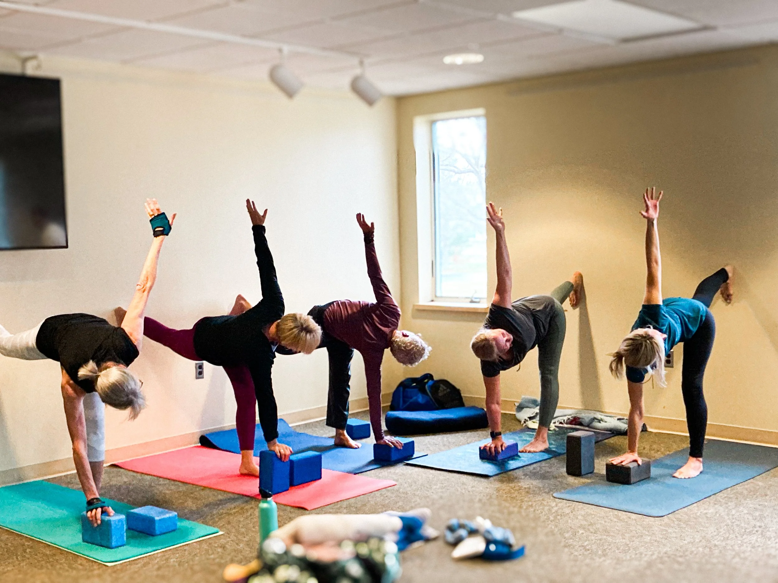 People practicing yoga in a room, performing a pose on yoga mats with blocks, focusing on balance and stretching.
