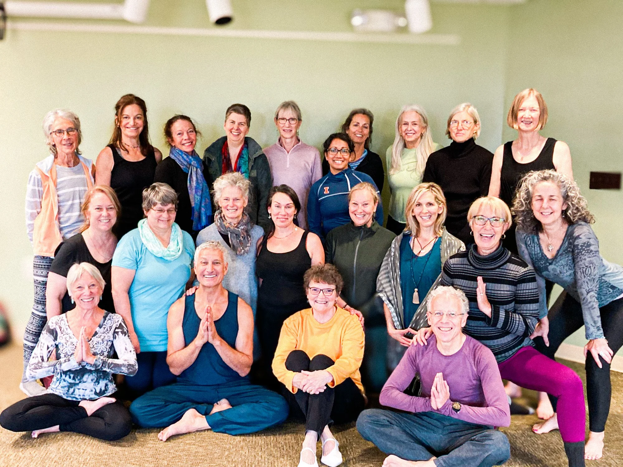 Group of women and a few men practicing yoga in a fitness or meditation class indoors, sitting cross-legged, some in prayer or meditation poses, smiling.