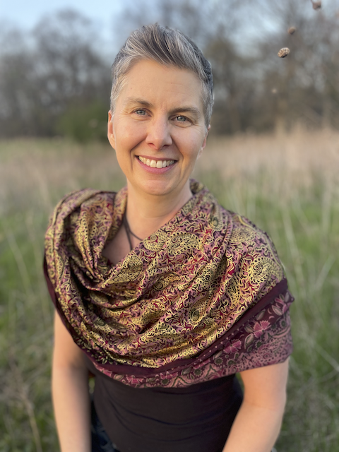 MYA Treasurer, Susan Freeman, with short hair standing outdoors in a field, wearing a patterned shawl over black clothing, with trees and sky in the background
