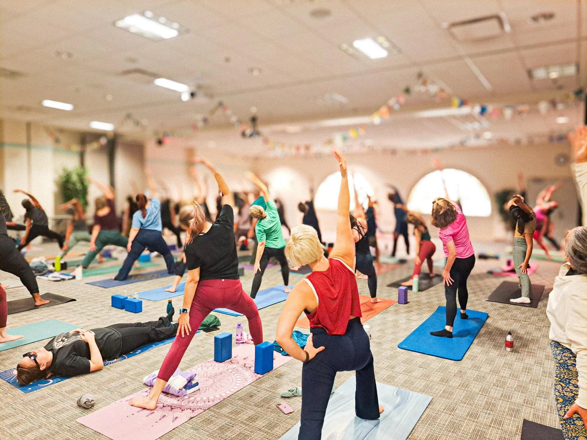 Group of people practicing yoga in a large indoor room with windows, some lying on mats and others in standing poses, with an instructor guiding the session.