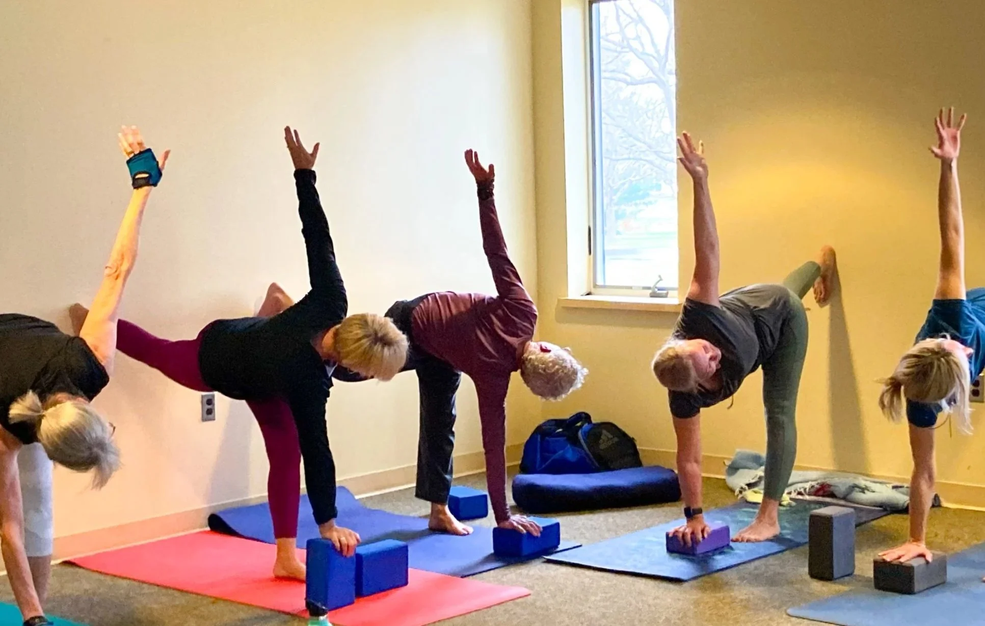 A group of older adults participating in a yoga or stretching class indoors, performing side stretches while balancing on one hand and foot on yoga mats, with blocks and backpacks nearby.