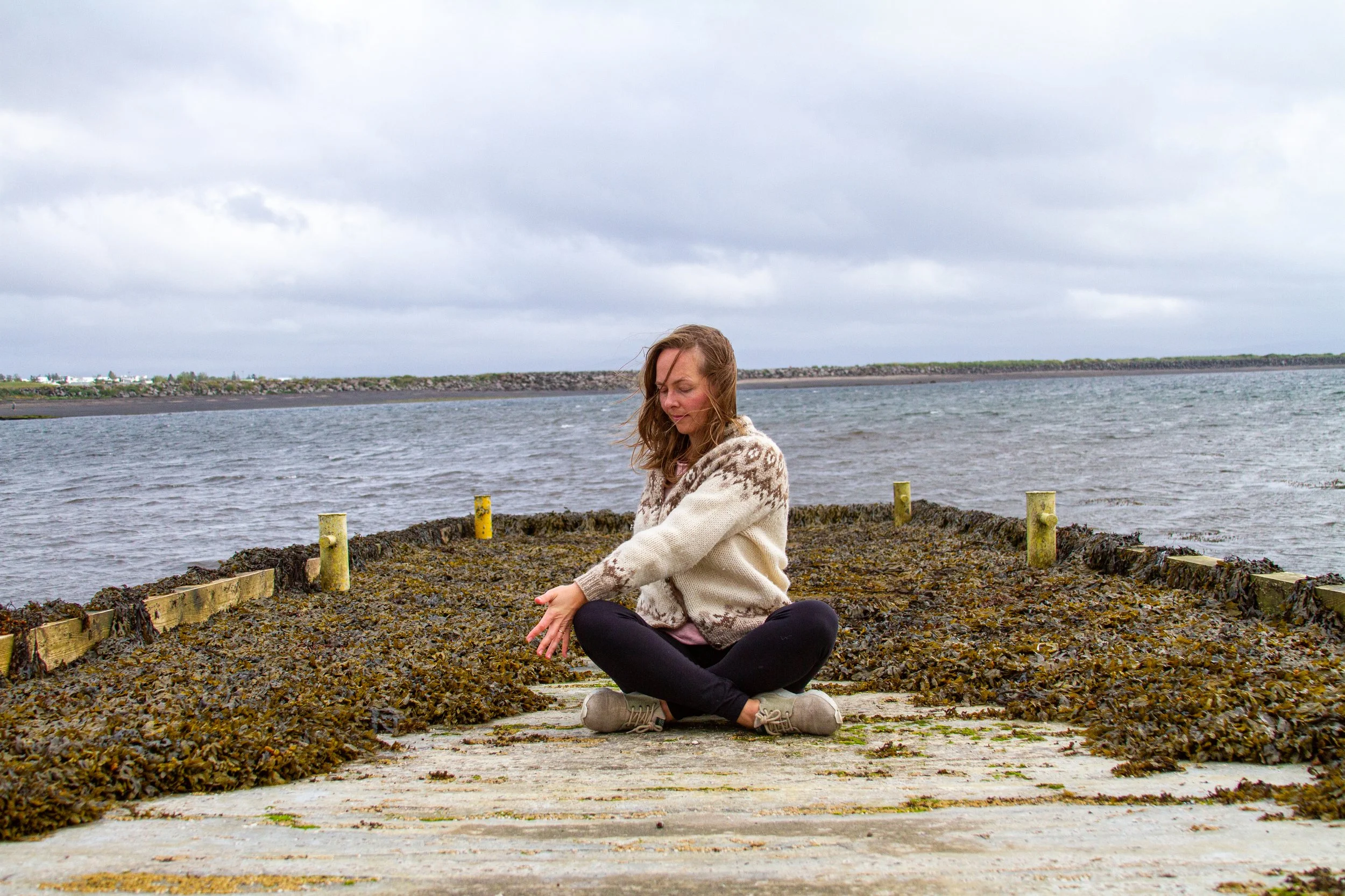A woman, Kristin Kristjansdottir, sitting cross-legged on a pier covered in seaweed, near water, with cloudy sky overhead.