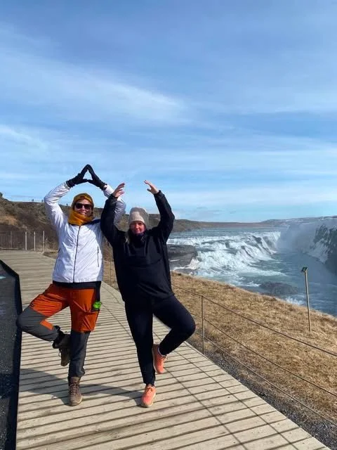 Two people making a heart shape with their arms beside an Icelandic waterfall.