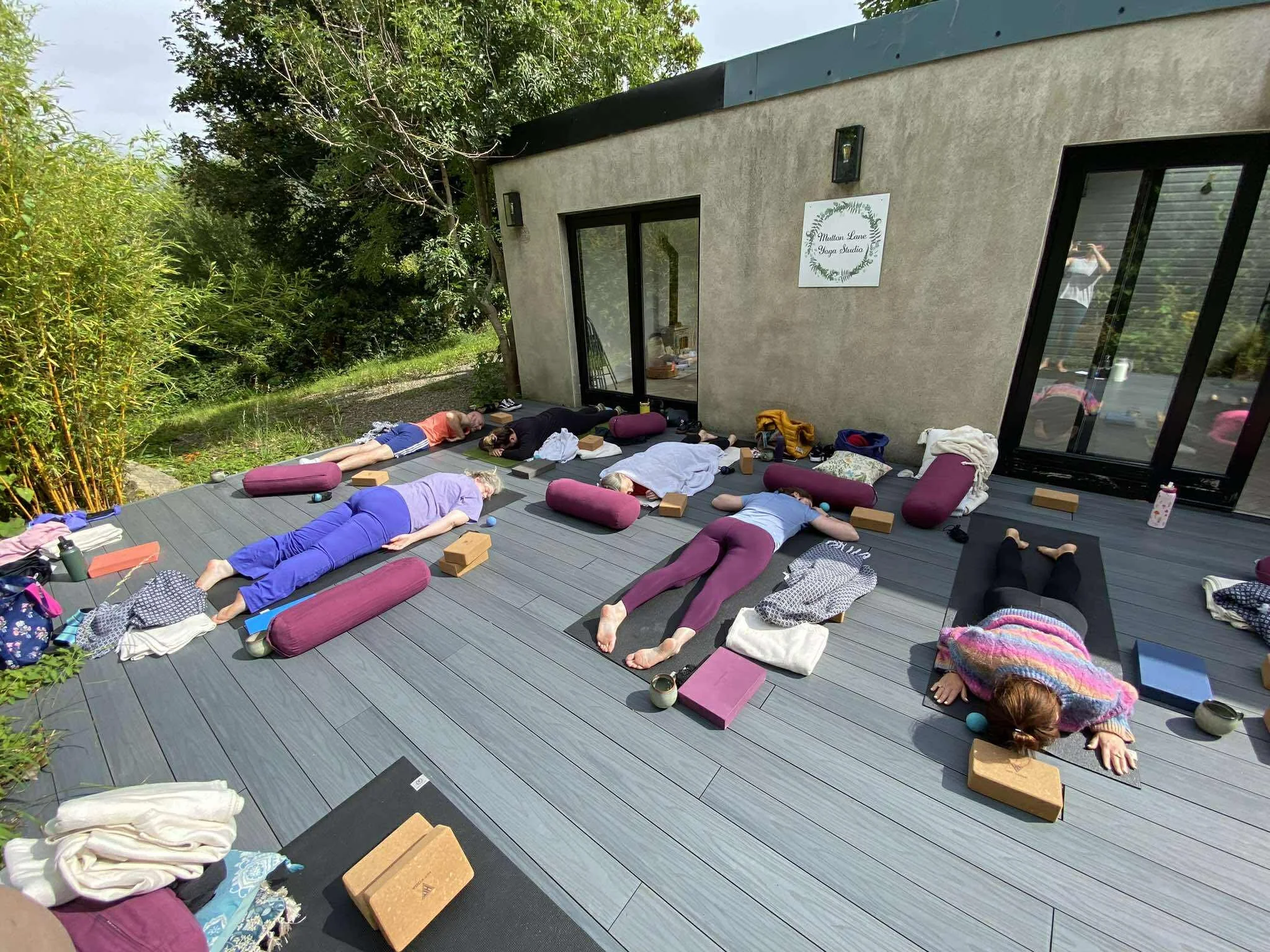 Group practising yoga outdoors on a wooden deck surrounded by greenery.