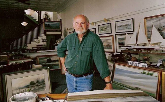 A man with a beard and bald head wearing a green shirt and jeans standing in an art gallery or store filled with framed paintings and model sailboats displayed on shelves and tables.