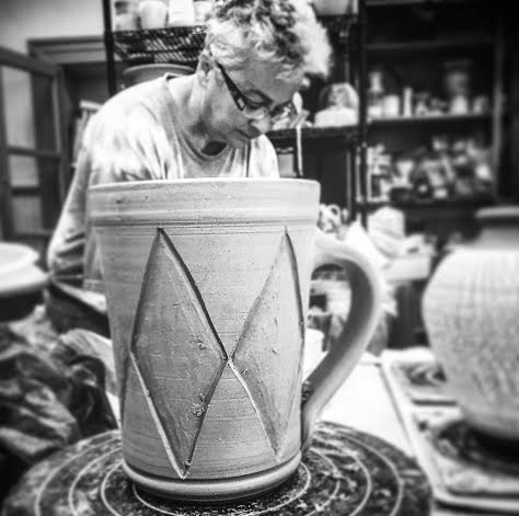 Black and white photo of an elderly woman working on a large ceramic mug with geometric designs in a pottery studio.