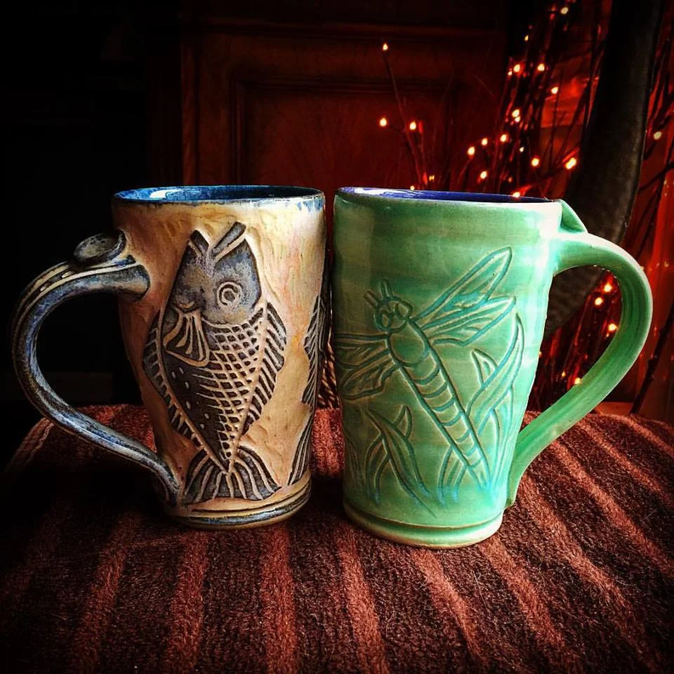 Two ceramic mugs with nature-themed designs, one with a carved owl and the other with a carved dragonfly, placed on a textured brown fabric surface with a dark background and decorative lights.