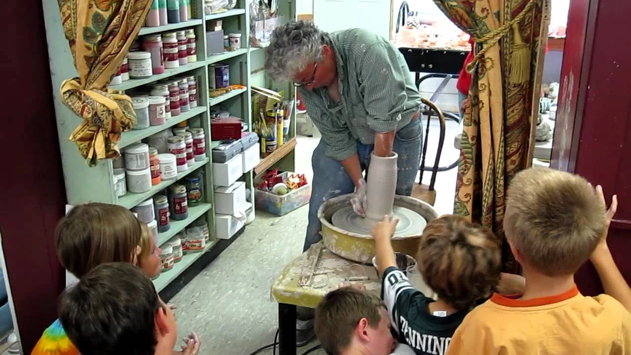 A woman demonstrating pottery on a pottery wheel while children watch in a room with shelves of supplies.