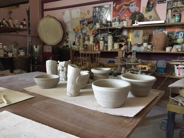 Studio workspace with unglazed ceramic bowls and vases on a worktable, surrounded by pottery tools, shelves with supplies, and artwork in the background.