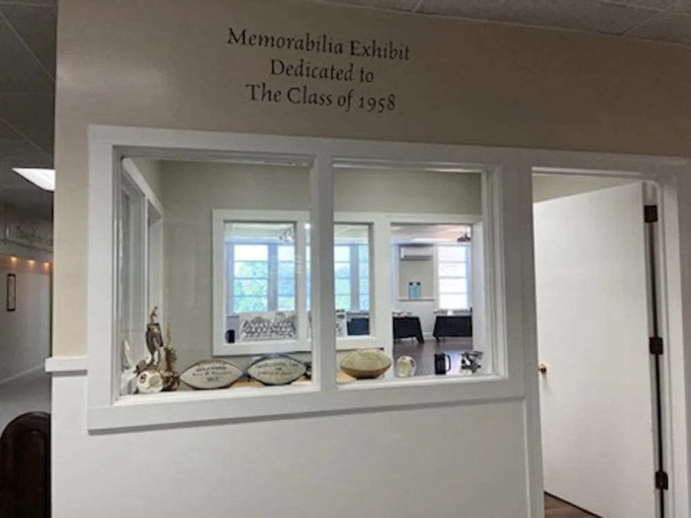 A sign on a wall reads 'Memorabilia Exhibit Dedicated to The Class of 1958'. Below the sign, a cut-out window shows a room with a table, chairs, and various memorabilia items.