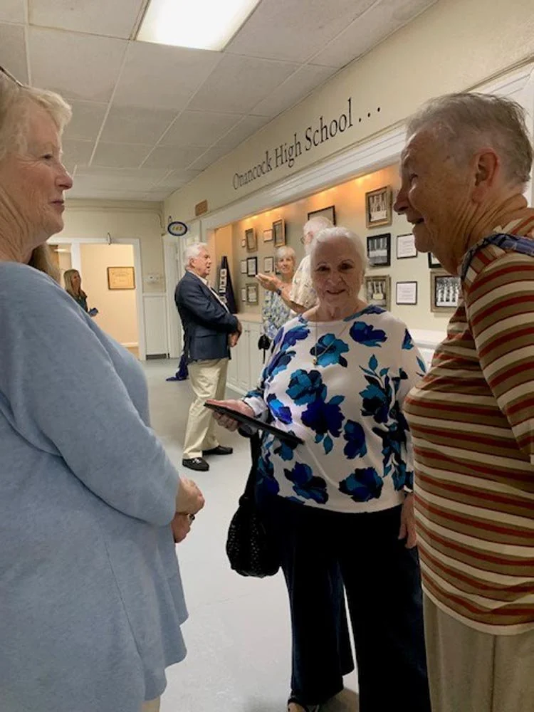Group of elderly people standing and talking in a hallway at Clemock High School, with framed photographs on the wall.