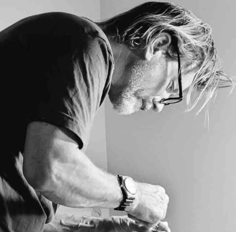 A man with glasses, a watch, and a beard, looking down and working on something, with short hair and wearing a dark shirt, in black and white.