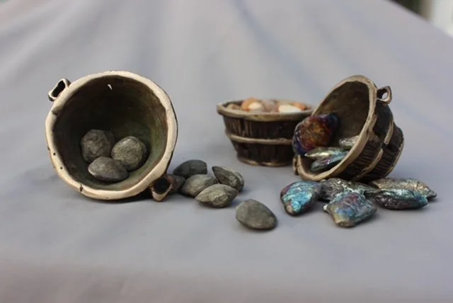 Small bowls filled with rocks and iridescent gemstones, with some rocks scattered on a gray cloth background.