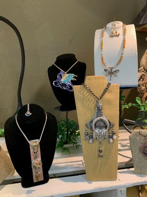 Display of various necklaces and jewelry on black and beige necklace busts, arranged on a table with decorative plants and objects.