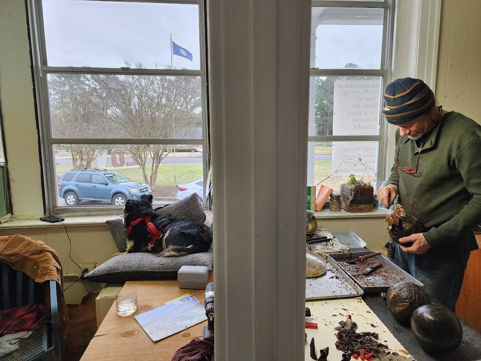 A split view of two rooms. The left side shows a room with a window, outside view of parked cars, a blanket-covered cushion with a dog lying on it, a dining table with papers, glasses, and clothing. The right side shows a man in a green hoodie and st
