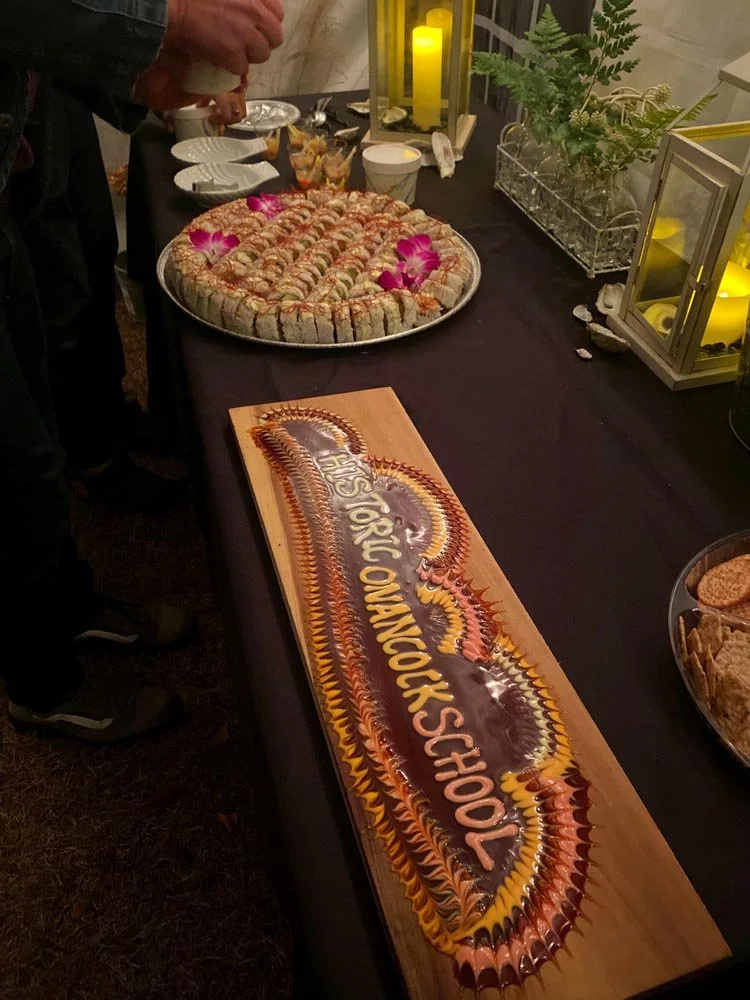 A buffet table with a large tray of sushi rolls decorated with pink flowers, a wooden sign with 'HS TOKYO ONGOLE SCHOOL' written in colorful lettering, and candles and greenery as decor.