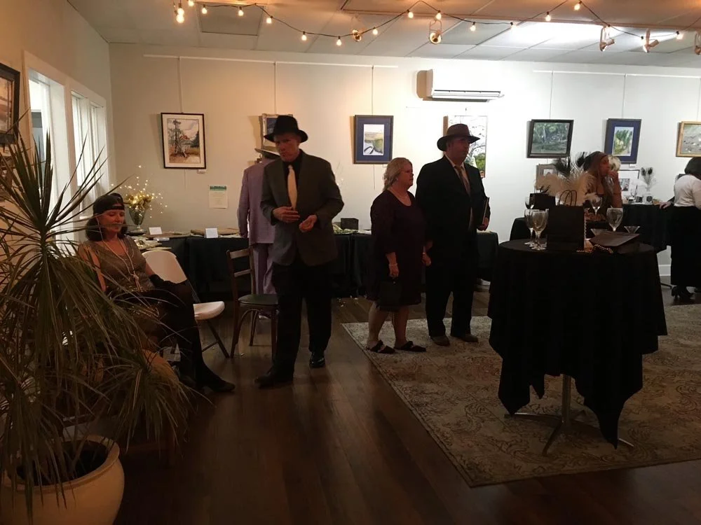 People gathered in an art gallery or exhibition space decorated with string lights. Some individuals are standing and chatting, while others are seated. Artwork hangs on the walls, and tables with drinks and decorations are present.