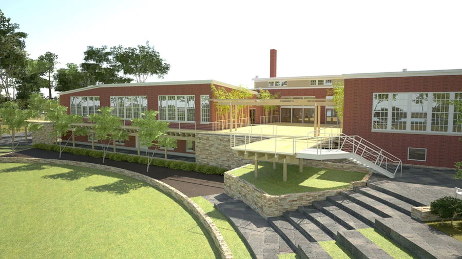 A modern, two-story brick building with large glass windows, surrounded by lush green trees, with a multi-level outdoor patio area with steps and railings.