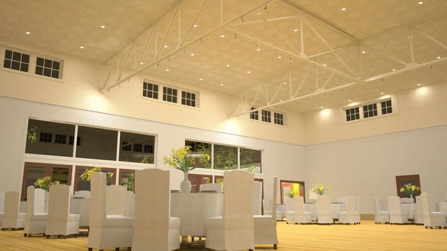 A spacious, well-lit event hall with high ceilings, white walls, and large windows. The room has white chairs and round tables decorated with vases of yellow flowers, likely set up for a gathering or celebration.