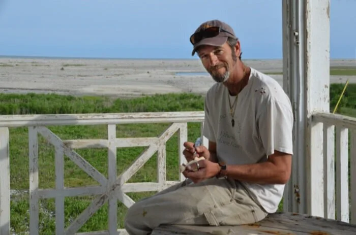 A man with a beard, wearing a cap and casual clothes, sitting on a porch near a beach, holding a bowl or container.