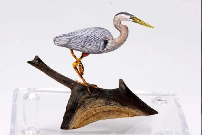 A mixed media art piece combining a heron with a bird's body, perched on a wooden branch displayed on a clear stand.
