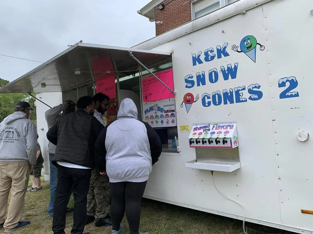 People standing in line at a snow cone stand with a sign that reads "KEK SNOW CONES 2" and a colorful display of shaved ice flavors.