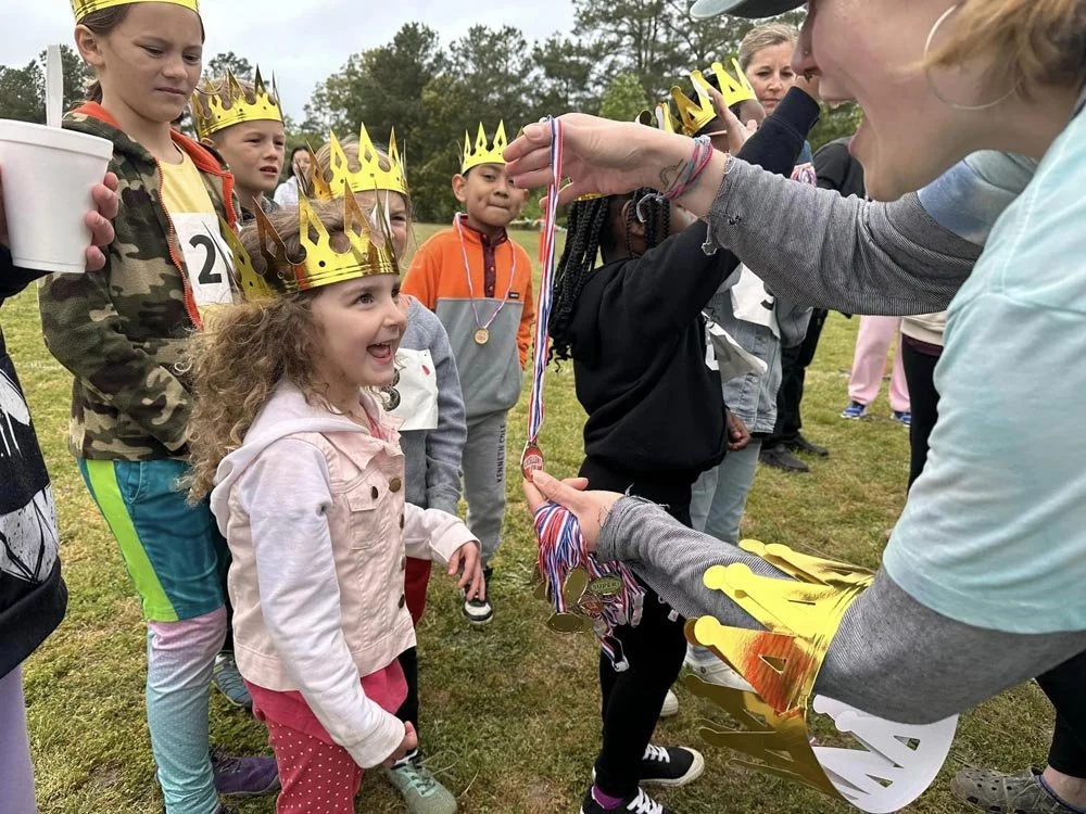 Children wearing paper crowns receive medals at an outdoor event, with kids standing in line on grass.