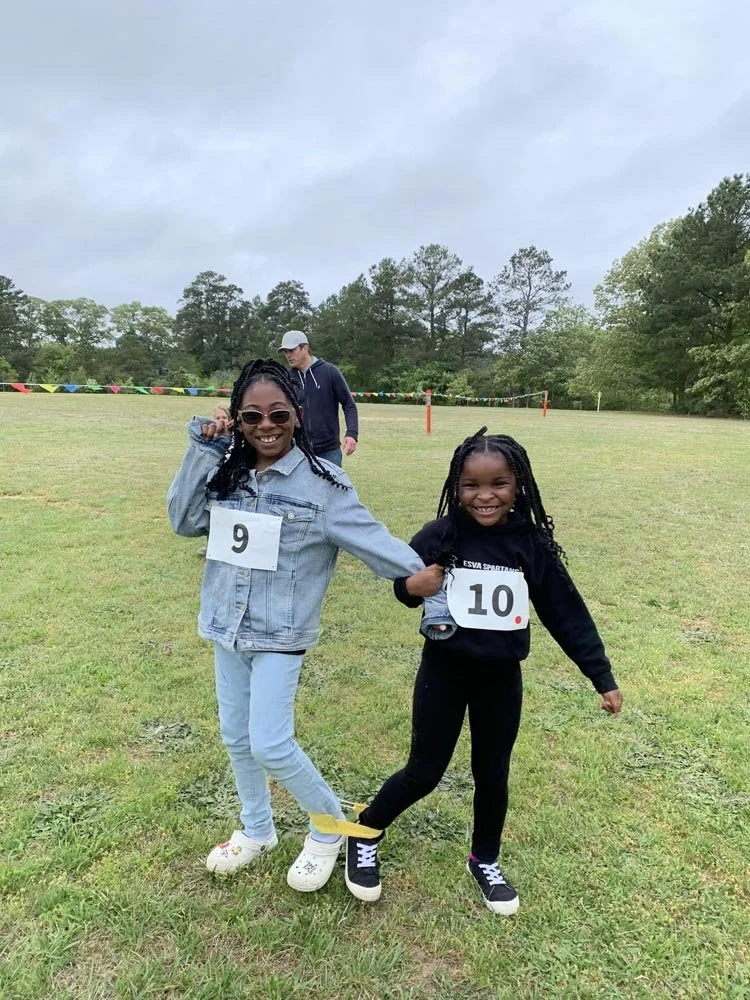 Two young girls, one wearing a jean jacket and the other in black, smiling and holding hands while participating in an outdoor race or event on a grassy field. They have race numbers 9 and 10 attached to their outfits.