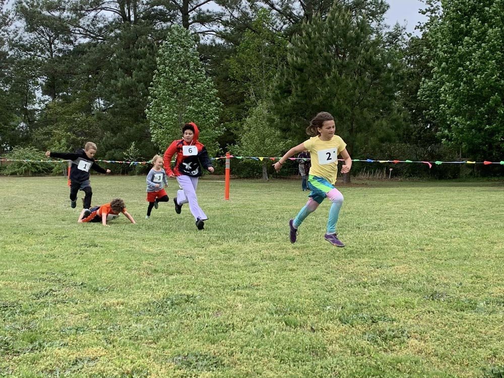 Children participating in a race on a grassy field, with some falling and others running towards the finish line, trees in the background.