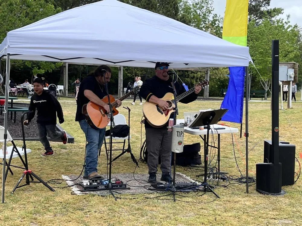 Two men playing acoustic guitars and singing on a small outdoor stage under a white canopy, with a man in the background running and a child nearby, during a outdoor event with trees and people in the background.