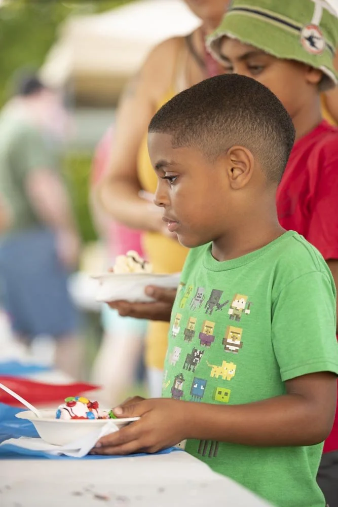 A young boy with a buzz cut wearing a green T-shirt with pixelated characters, holding a bowl of ice cream, at a gathering or outdoor event.