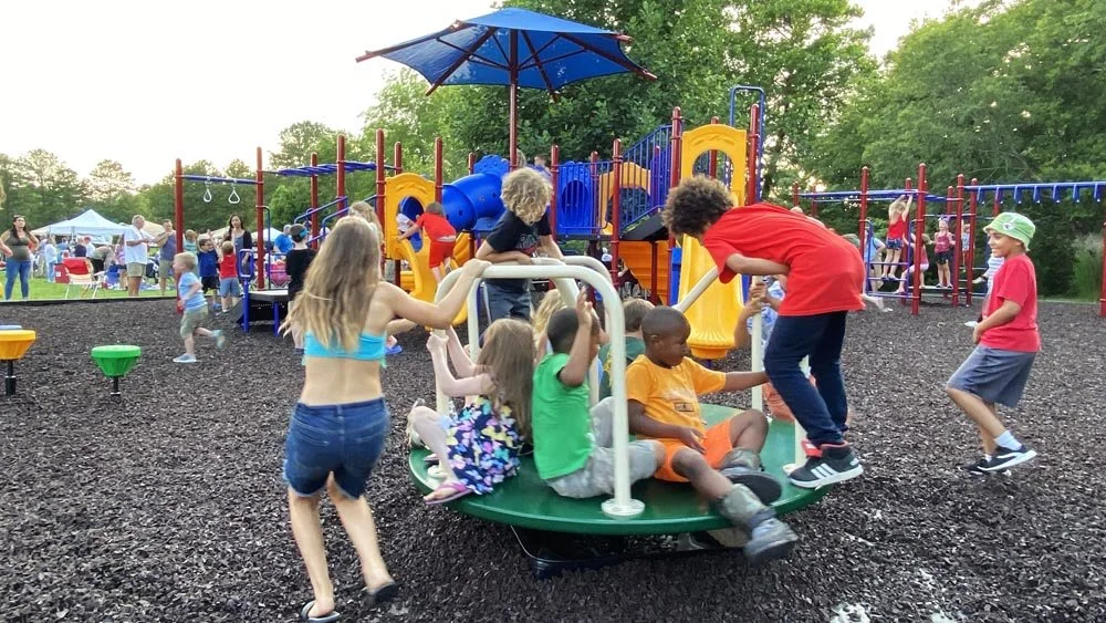 Children playing on a playground at a park with a colorful structure, slides, and a large umbrella for shade, with adults and other kids in the background enjoying the outdoor space.